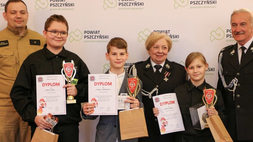 Powiększ: Dzieci trzymające dyplomy i statuetki oraz dorośli w mundurach strażackich.