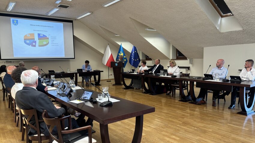 Powiększ: Osoby siedzące przy stołach, przy mównicy stoi kobieta, w tle na ekranie wyświetla się prezentacja. 