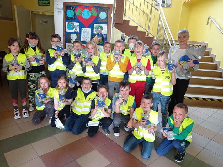 Powiększ: Na zdjęciu grupa uśmiechnietych dzieci w odblaskowych kamizelkach trzymających w ręku maskotkę obok dzici z prawej strony dyrektorka szkoły